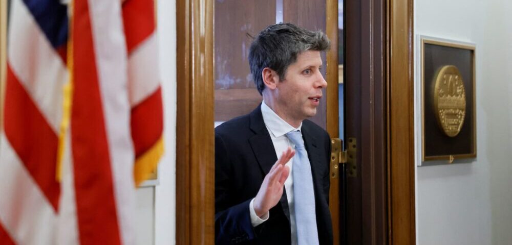 Article Image - Best AI Tools Man in a dark suit and blue tie steps through a doorway, with an American flag visible on the left.