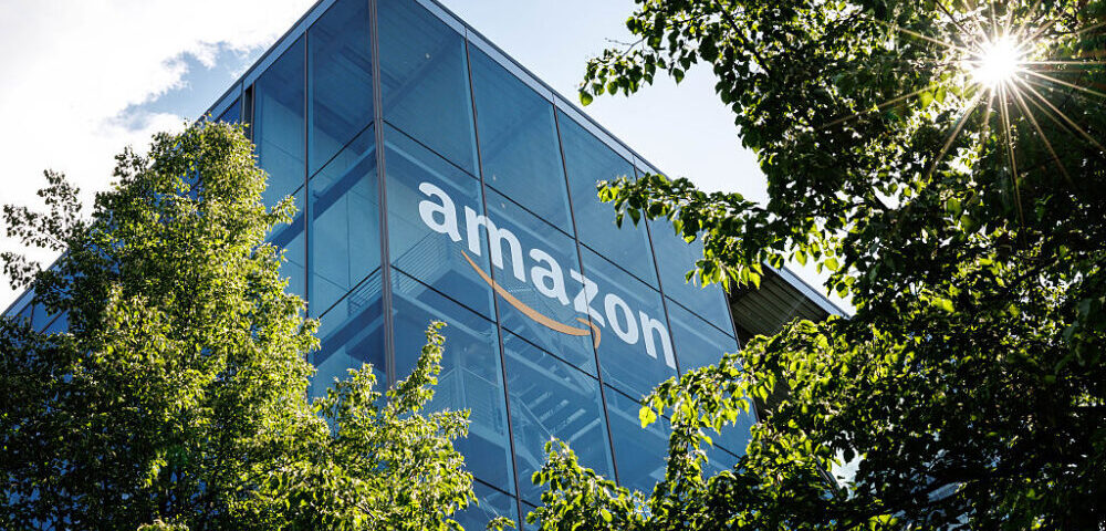 Article Image - Best AI Tools Amazon logo on a modern glass office building surrounded by green trees and bright sunlight.