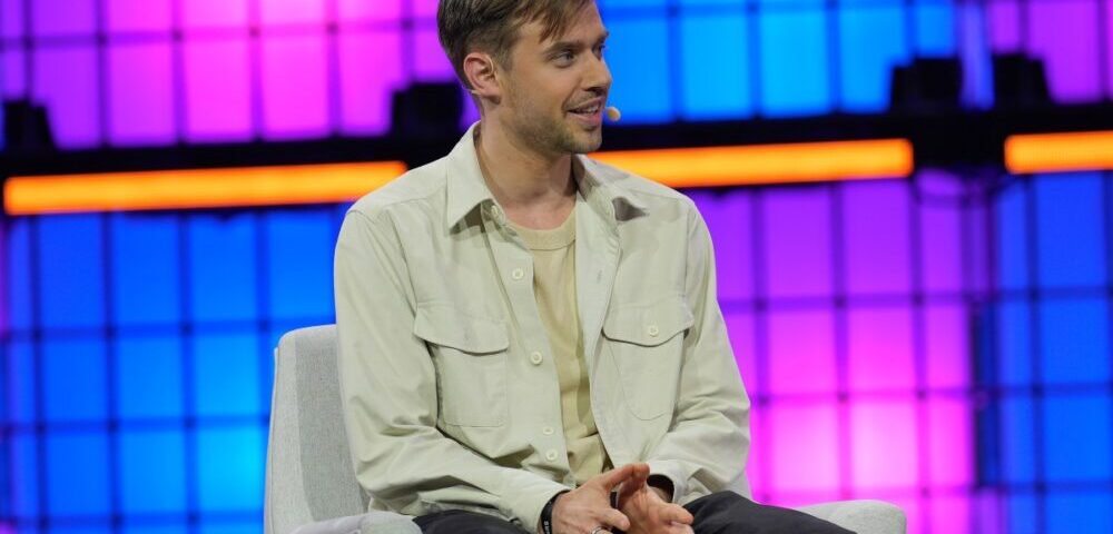 Article Image - Best AI Tools Man in a light jacket with a headset microphone sits on a chair on a brightly lit stage with purple and blue panels in the background, speaking or answering questions.