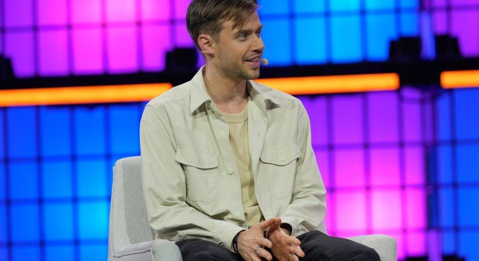 Article Image - Best AI Tools Man in a light jacket with a headset microphone sits on a chair on a brightly lit stage with purple and blue panels in the background, speaking or answering questions.