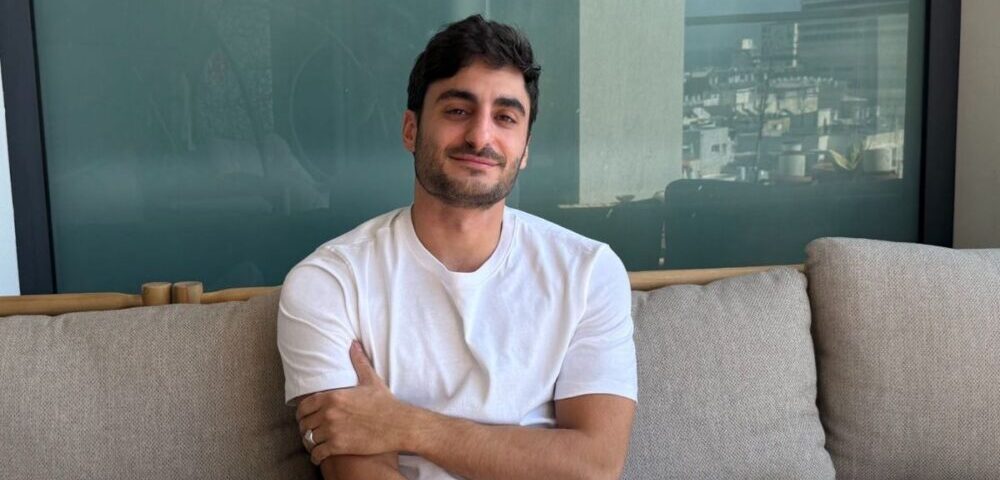 Article Image - Best AI Tools Man with dark hair and beard sits on a beige couch, smiling at the camera, wearing a white T-shirt.