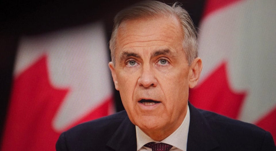 Middle-aged man in a dark suit and tie speaking, with blurred Canadian flags in the background.