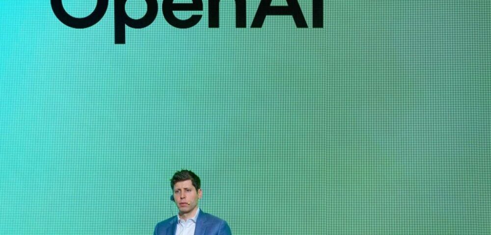 Article Image - Best AI Tools Man in a blue suit sits on stage against a green OpenAI backdrop with branding above him.