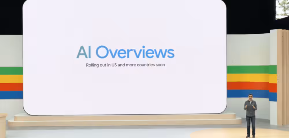Stage presentation with a large white screen showing 'AI Overviews' and subtitle 'Rolling out in US and more countries soon', while a speaker stands on the right.