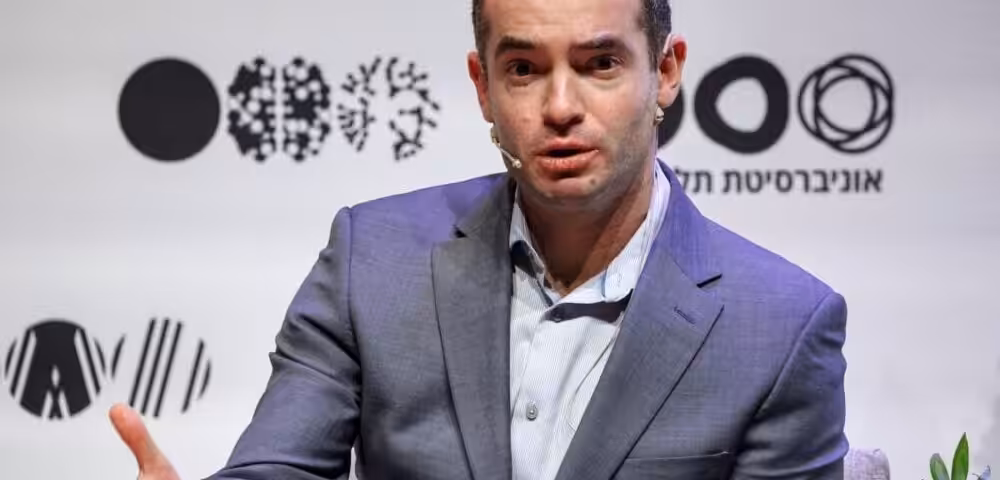 Male speaker in a gray suit addressing an audience on a panel, wearing a headset mic, with logos on a white backdrop behind him.