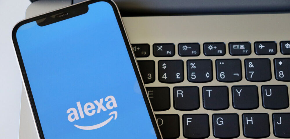 Article Image - Best AI Tools Smartphone showing the Alexa app logo on a blue screen beside a laptop keyboard, indicating voice assistant use on mobile.
