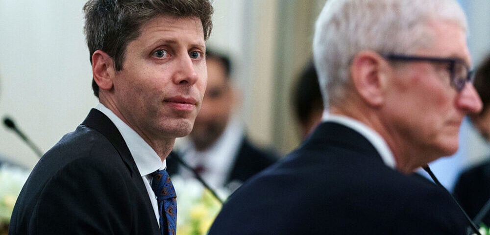 Article Image - Best AI Tools Man in a dark suit and blue patterned tie looks toward the camera at a formal event, with another suited man blurred in the foreground.