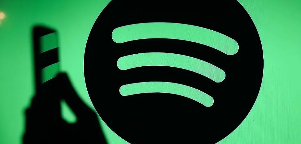 Article Image - Best AI Tools Close-up of the Spotify logo on a bright green background with a shadowy hand holding a striped object to the left