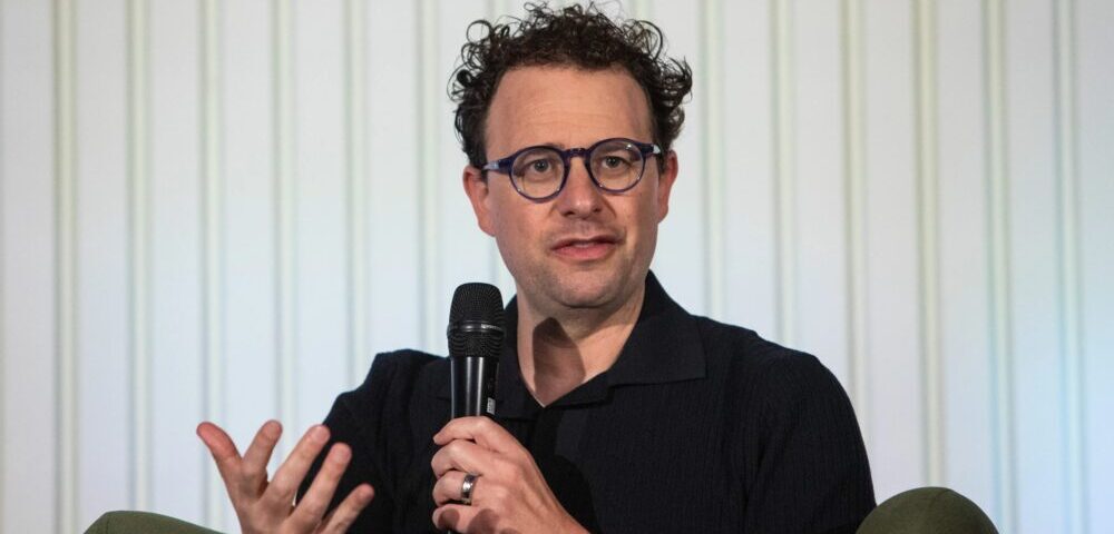 Article Image - Best AI Tools Man wearing blue-rimmed glasses speaks into a handheld microphone during a panel discussion.