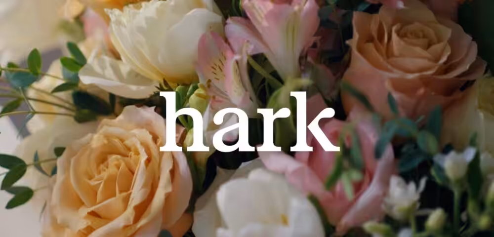 Bouquet of peach, pink, and white roses with green foliage and the word 'hark' overlaying the center as brand text.