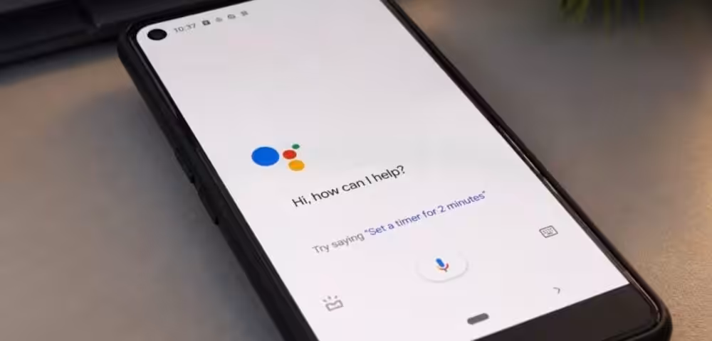 Smartphone on a desk showing Google Assistant welcome screen: ‘Hi, how can I help?’ with microphone button visible