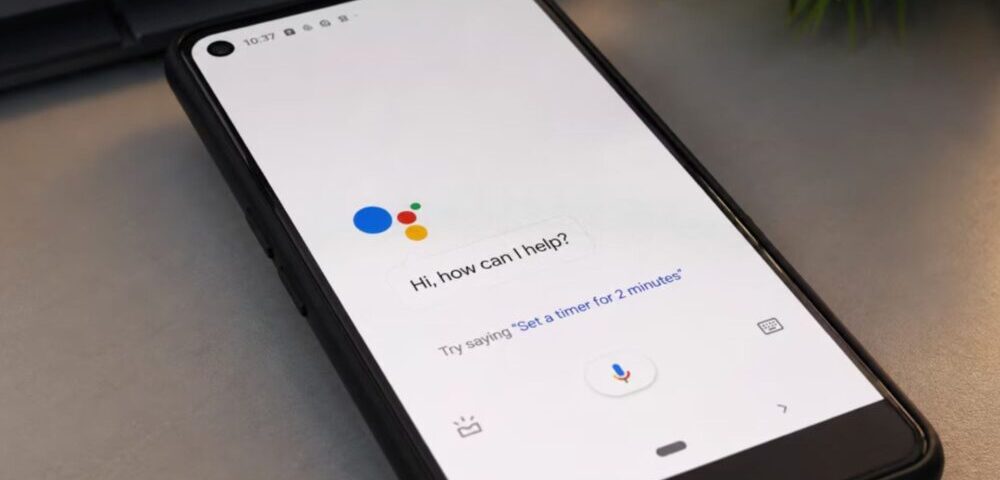 Article Image - Best AI Tools Smartphone on a desk showing Google Assistant welcome screen: ‘Hi, how can I help?’ with microphone button visible