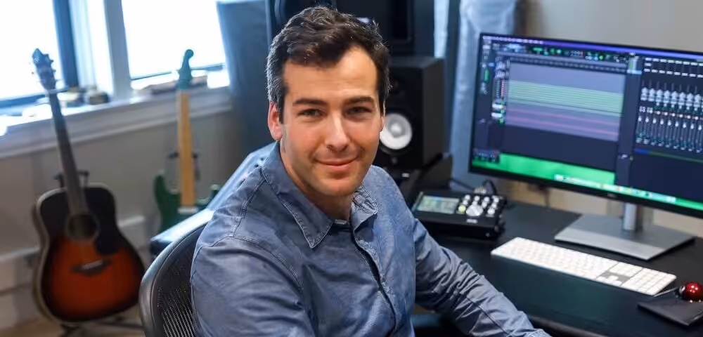 Man in a blue shirt sits at a home recording studio desk, with guitars in the background and a DAW on the monitor.