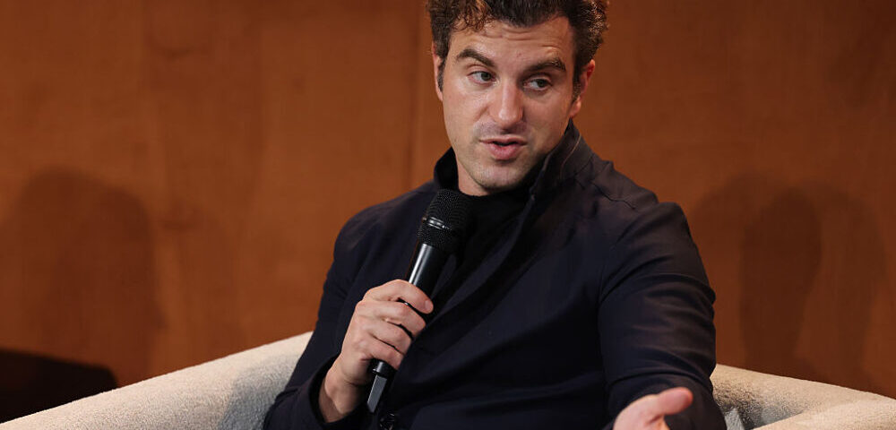 Article Image - Best AI Tools Man in a dark blazer holding a microphone, speaking and gesturing during a discussion on a panel.